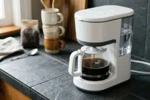 Cafetière filtre électrique blanche en fonctionnement sur comptoir ardoise gris, tasses empilées en arrière-plan
