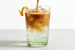 Verre d espresso tonic avec glacons, degrade brun ambre et zeste d orange, fond gris clair